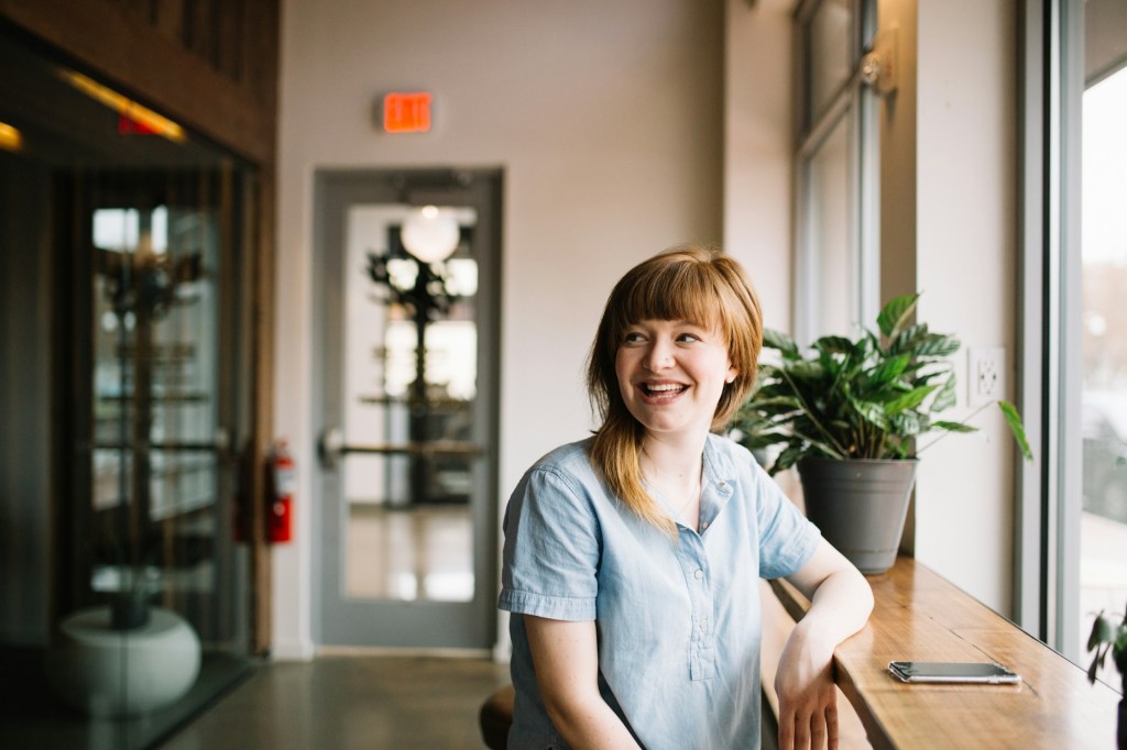 woman with reddish blonde hair and bangs wearing a pale-blue button-up shirt looking to the side and smiling