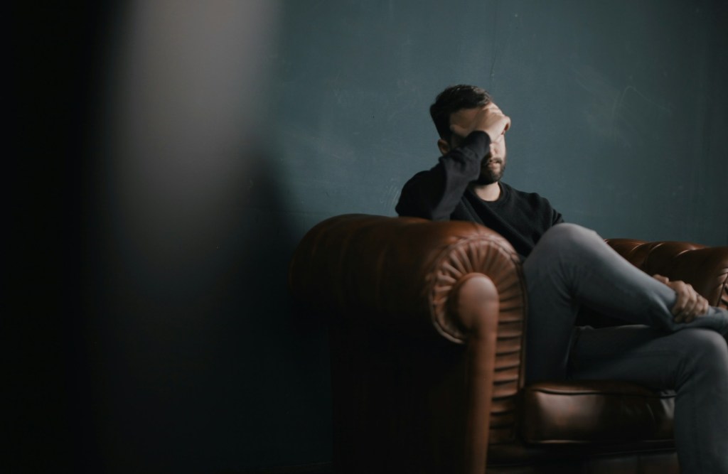 Man sitting with head in hands on a leather coach with blurred section to the left of the frame